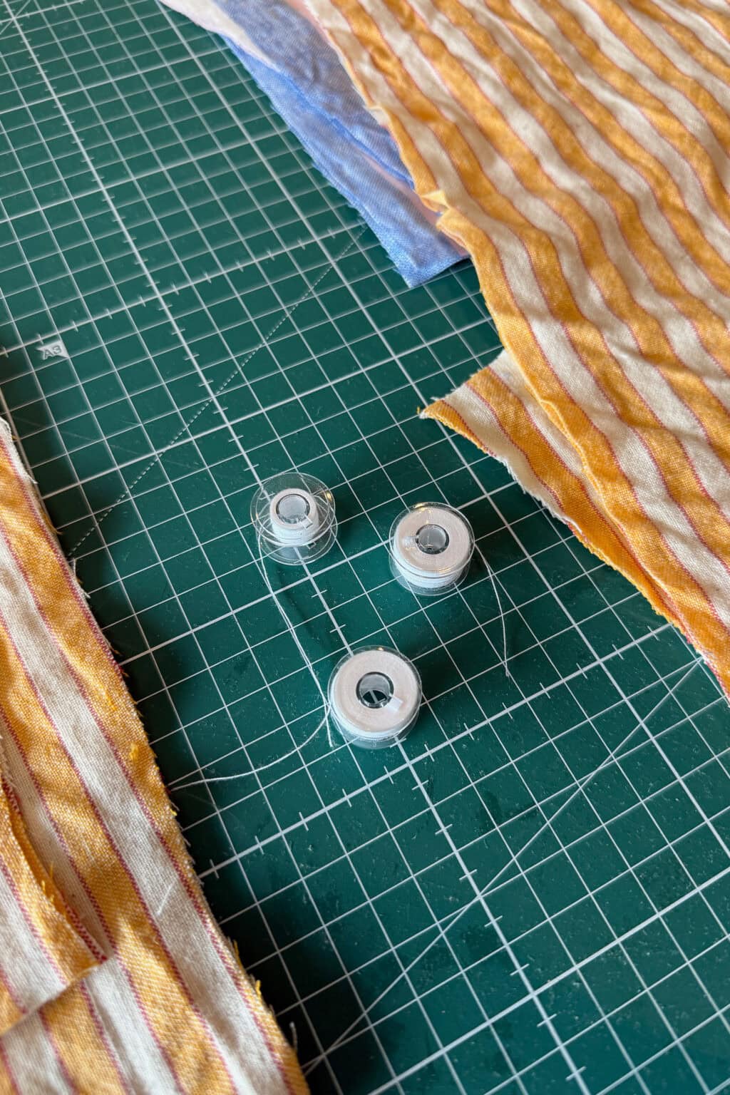 Three sewing machine bobbins and pieces of striped fabric in yellow and white are placed on a green cutting mat with a grid pattern.