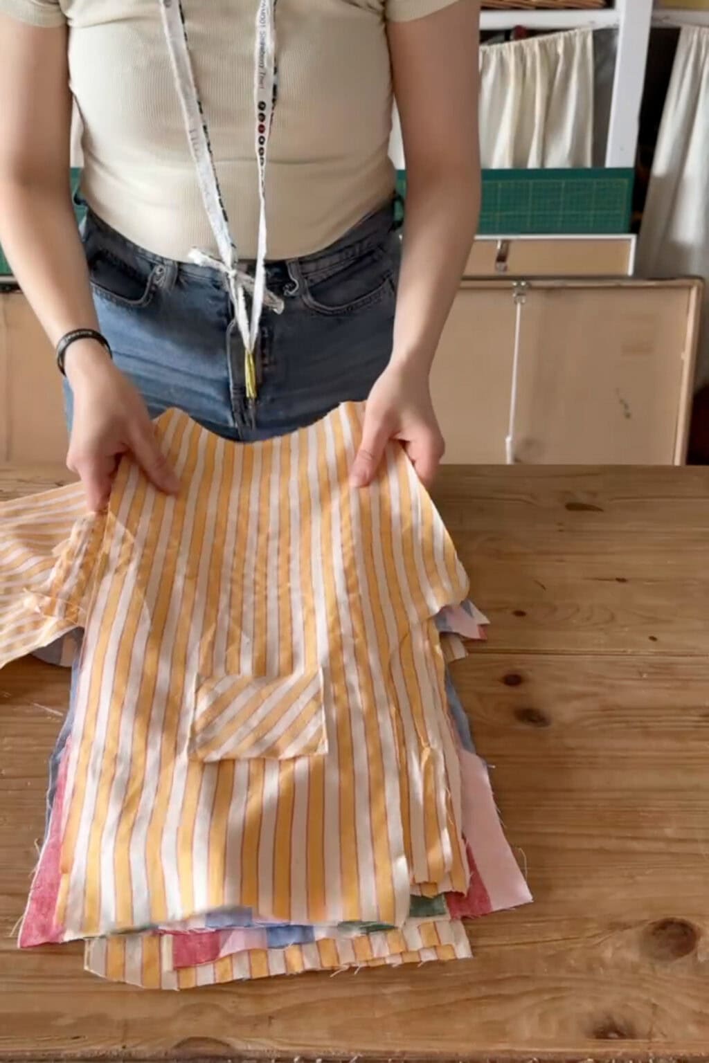 A person holds up a neatly folded stack of striped and colourful fabric pieces on a wooden table, with sewing tools and shelves in the background.