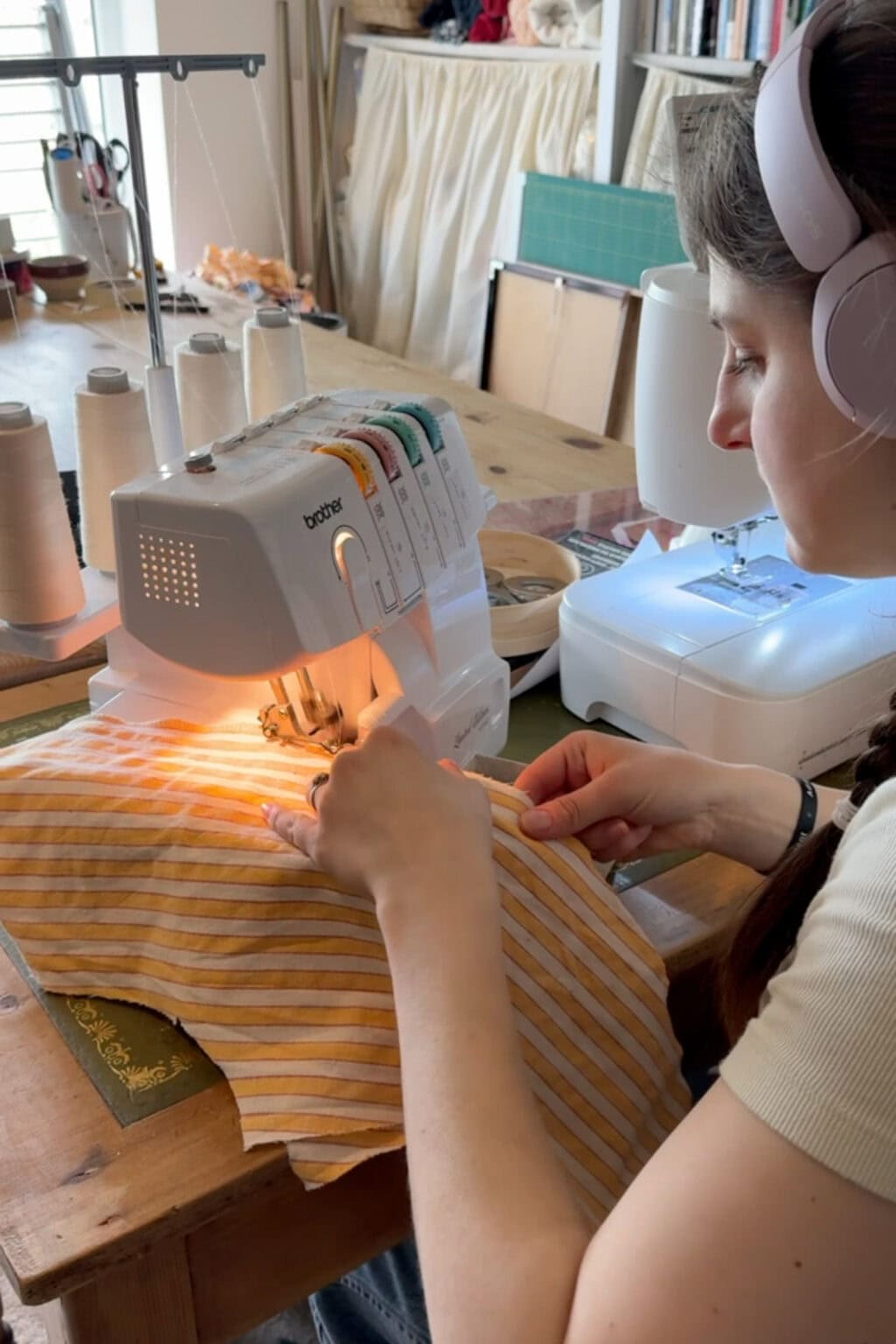 A woman wearing headphones uses a Brother overlocker sewing machine to stitch yellow and white striped fabric at a wooden table, with thread reels and another sewing machine nearby.
