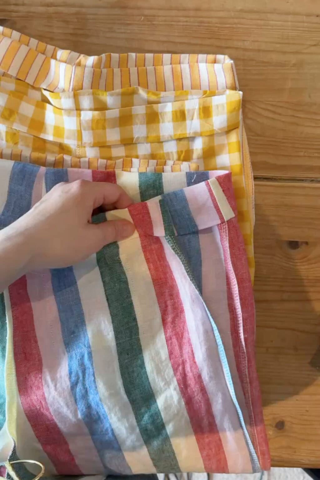 A hand holds the waistband of a pair of trousers with vertical red, blue, green, and white stripes. Beneath it is another pair with a yellow and white chequered pattern, both lying on a wooden surface.