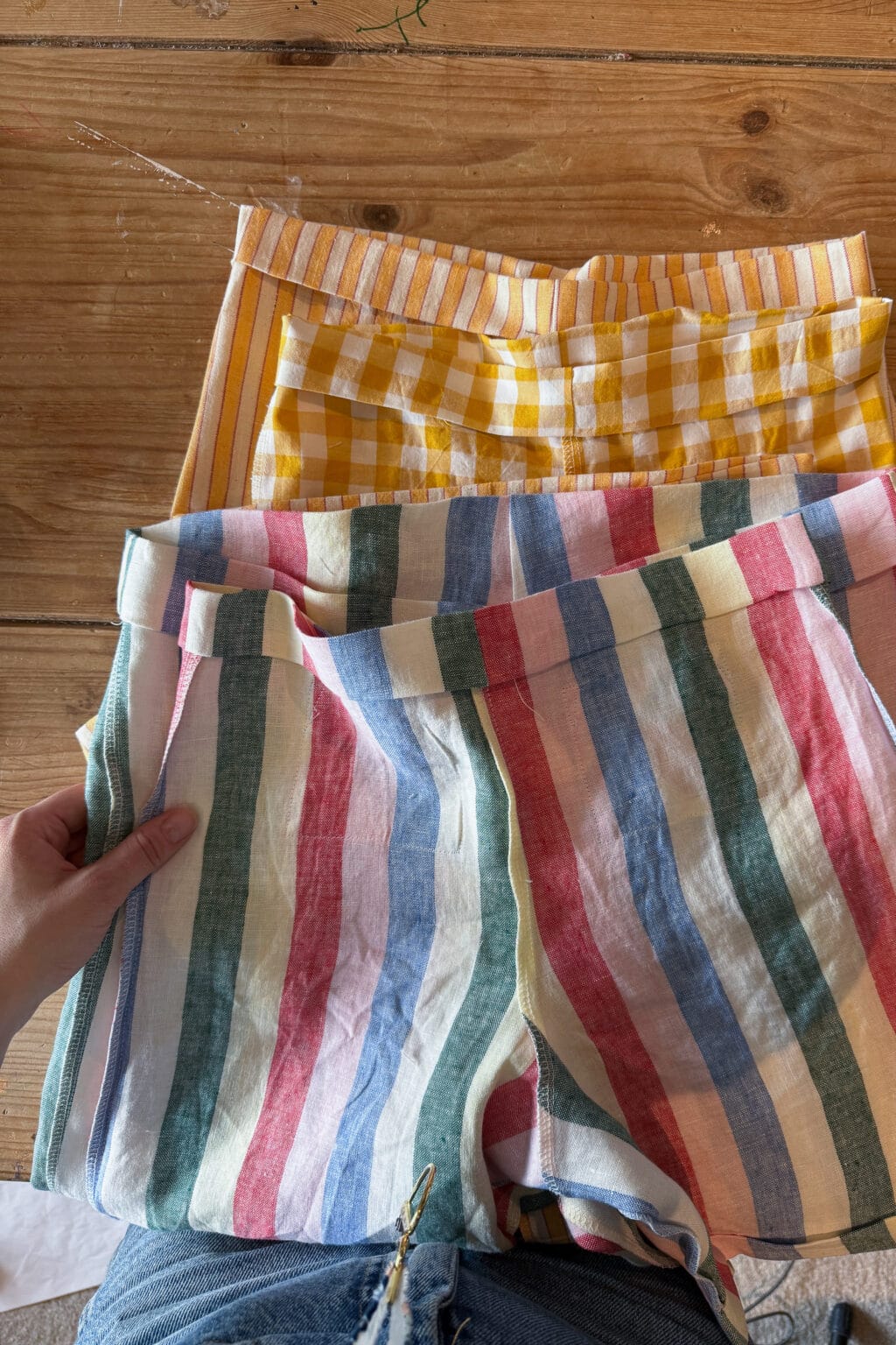 A hand holds a pair of pastel striped shorts over another pair of yellow gingham shorts, both laid on a wooden surface. The person is wearing blue denim shorts.