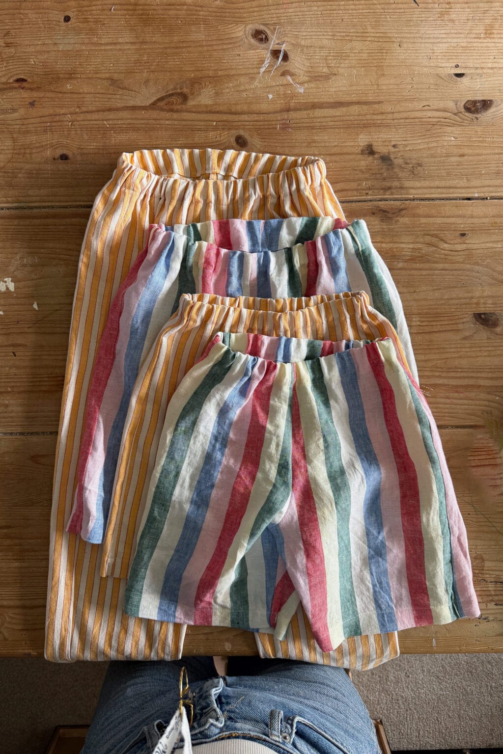Three pairs of elastic-waist shorts with vertical stripes, in different sizes, are laid out on a wooden table. The shorts have colourful stripes in yellow, orange, blue, green, and pink.
