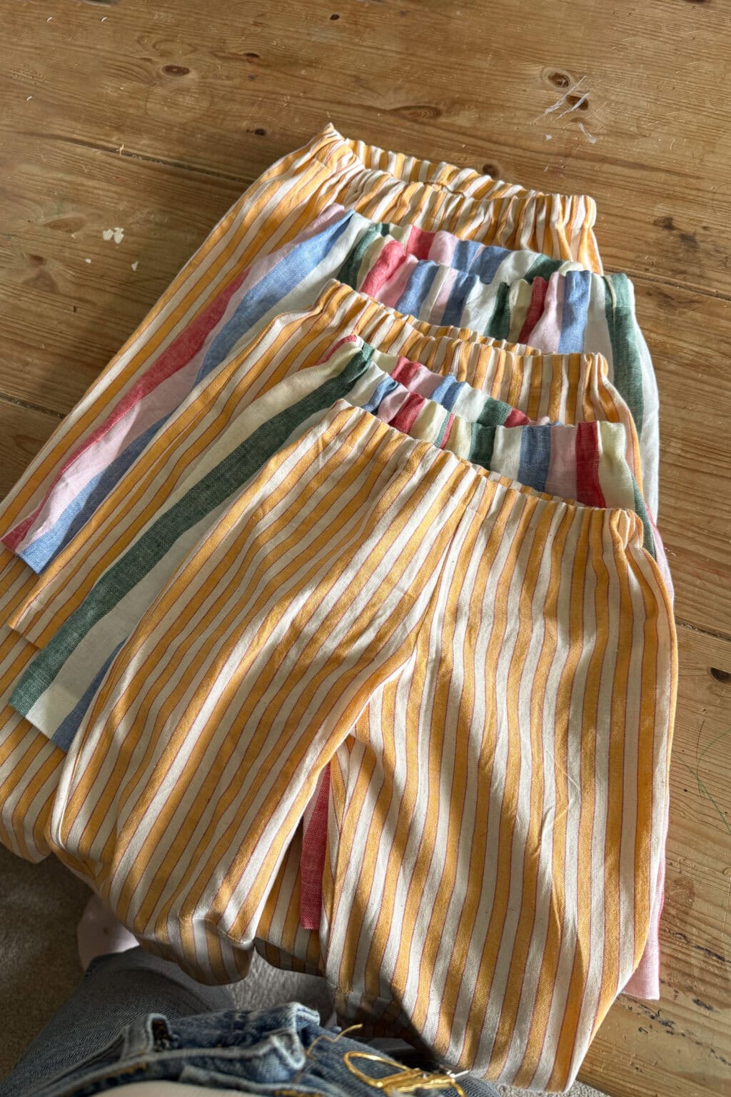 Four pairs of elastic-waisted shorts with striped patterns, two with yellow and white stripes and two with multicoloured stripes, are laid out on a wooden surface.
