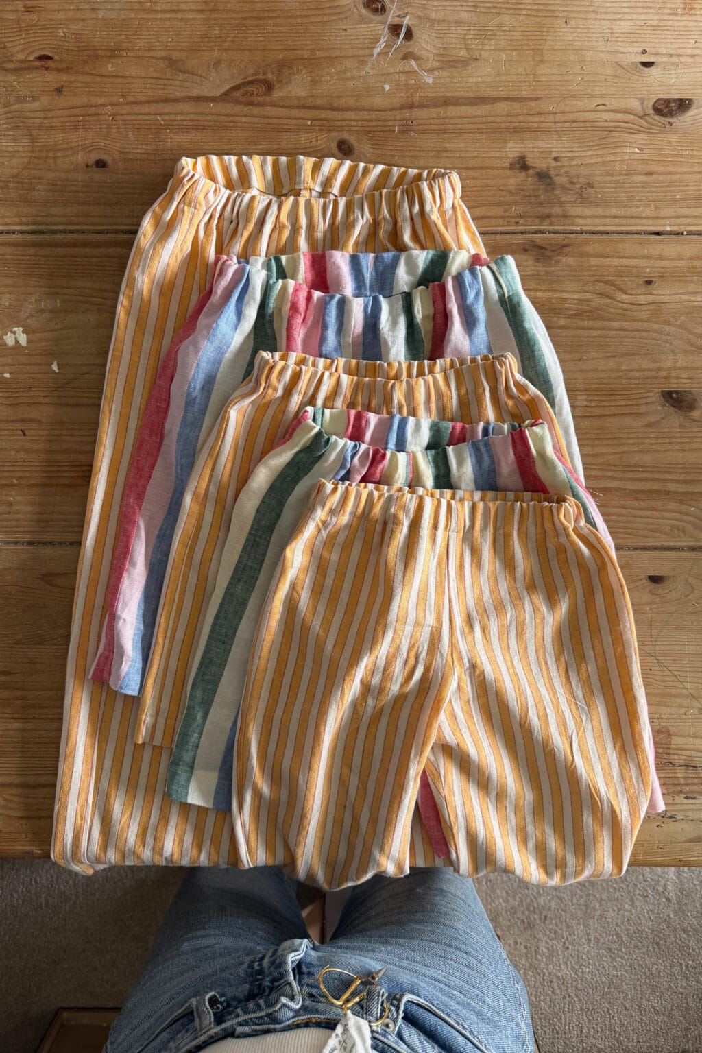 Four pairs of elastic-waist shorts in varying sizes are laid out on a wooden table. Two pairs have yellow and white stripes, whilst the other two have multicoloured vertical stripes. Blue jeans are visible at the bottom of the image.