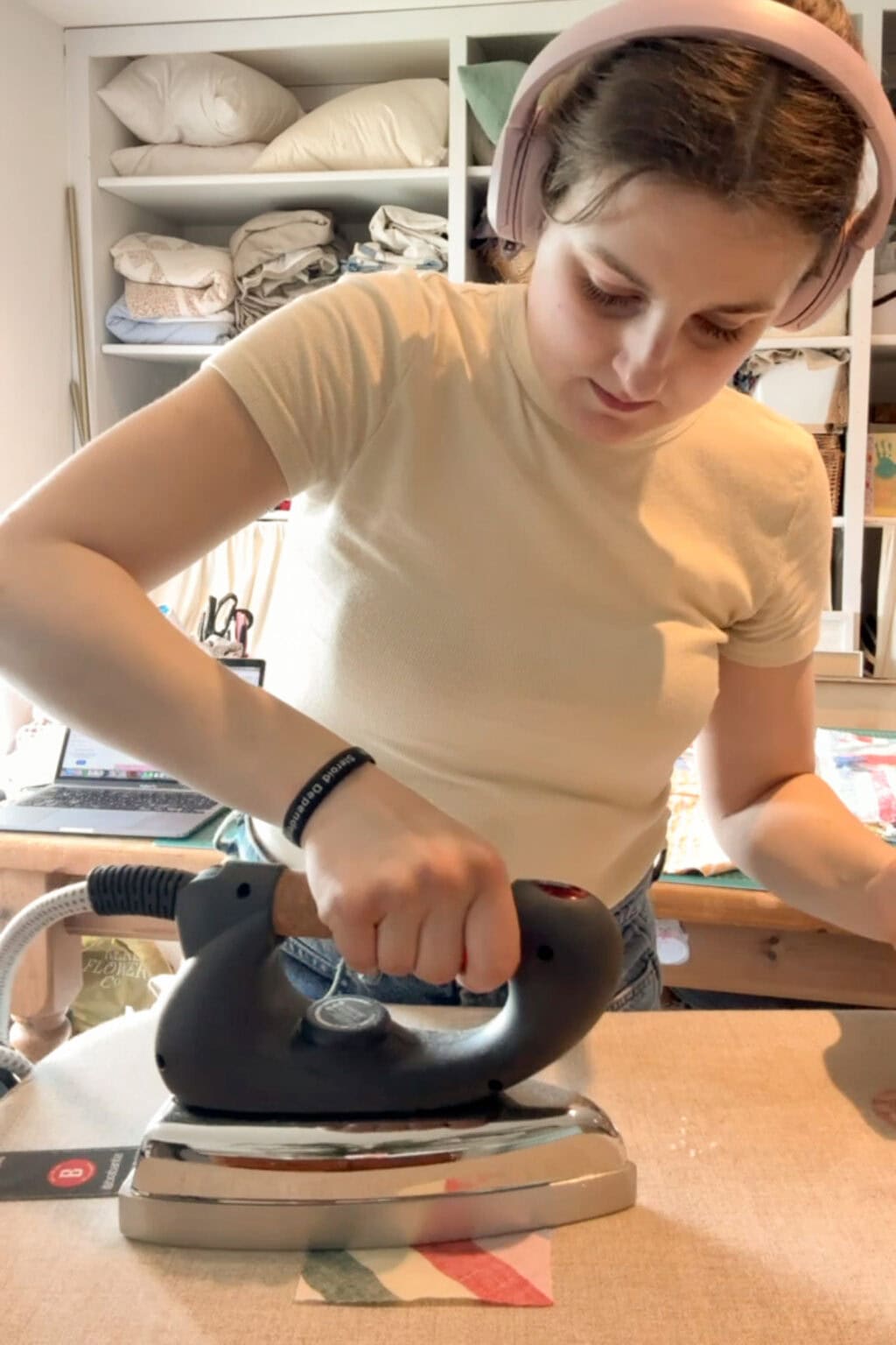 A woman wearing headphones and a light-coloured T-shirt is ironing a piece of fabric on a table in a cosy, organised room with shelves of folded linen and craft supplies in the background.