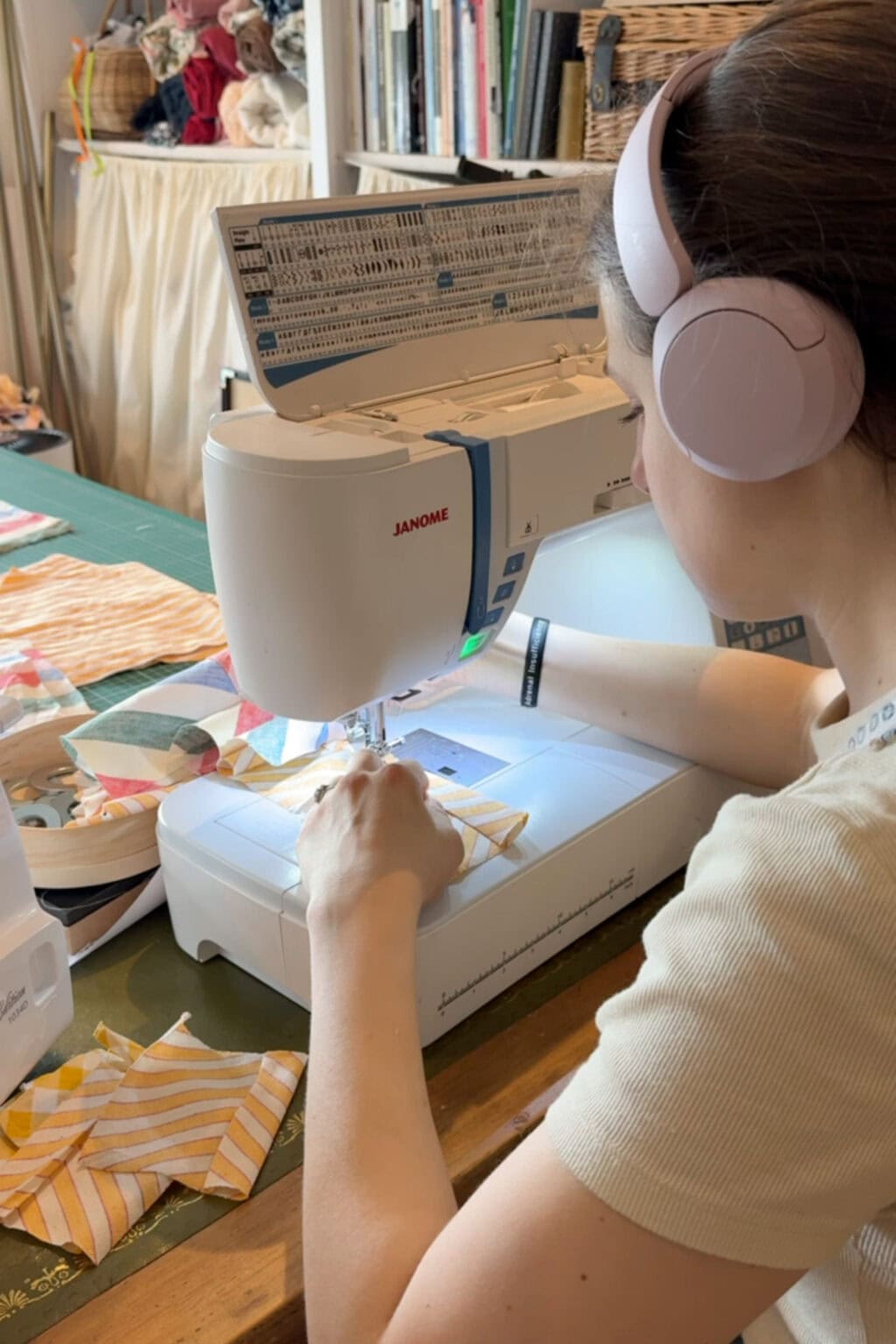 A person wearing pink headphones uses a Janome sewing machine to stitch pieces of fabric. Various fabric scraps are scattered on the workspace, and shelves with craft supplies are visible in the background.