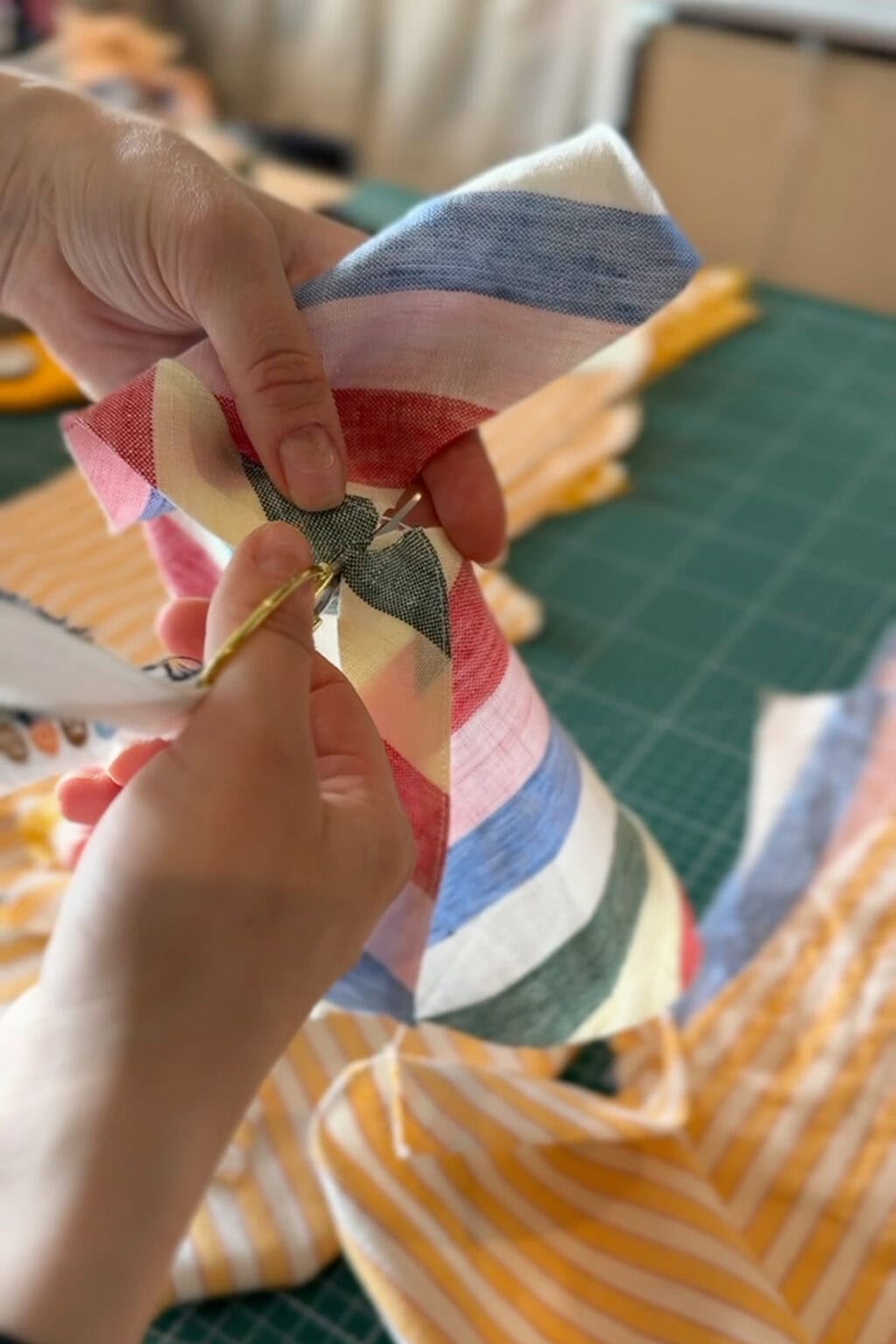 Close-up of hands using scissors to cut a piece of colourful, striped fabric on a green cutting mat, with yellow striped fabric and sewing tools visible in the background.