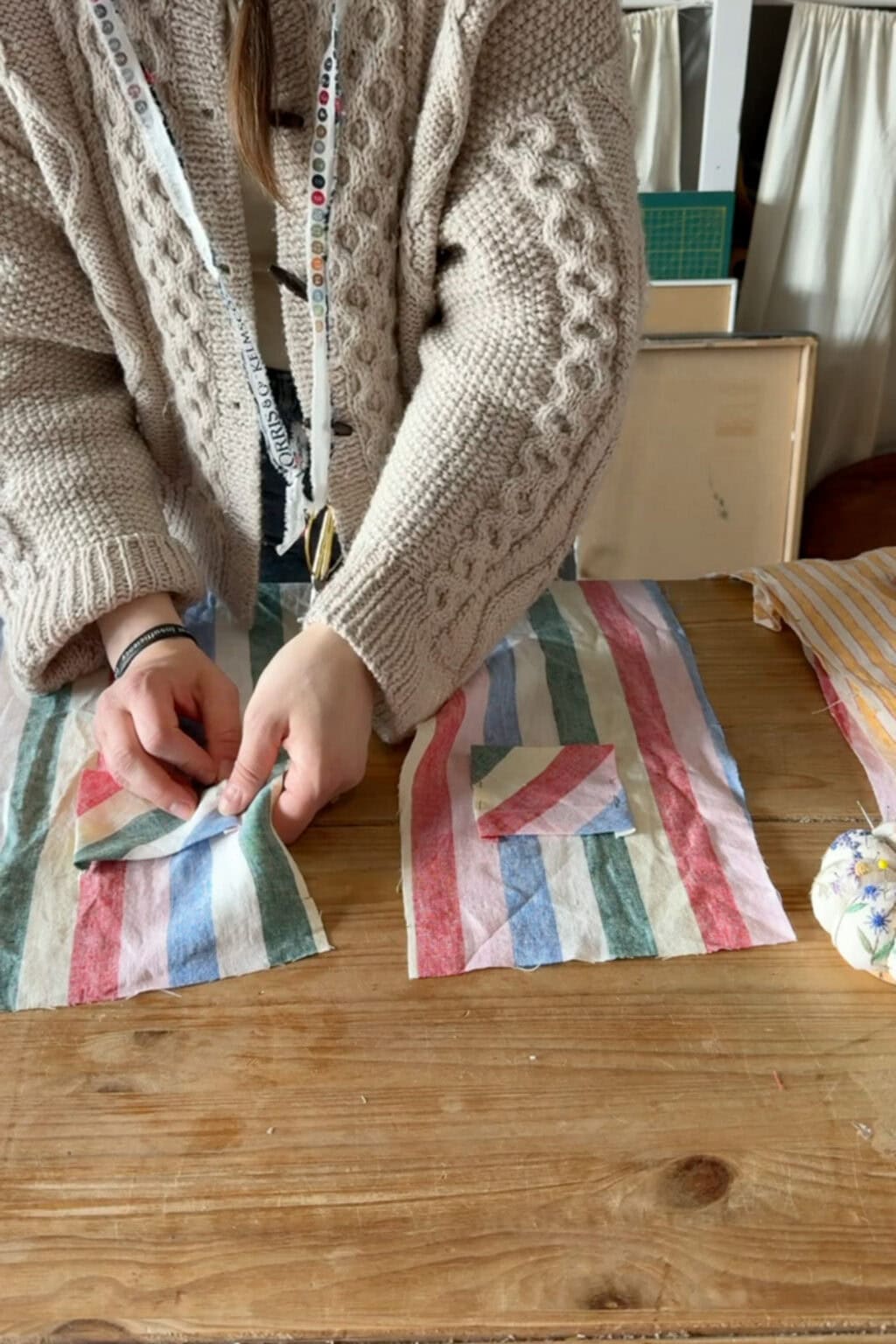 A person wearing a beige knitted jumper works with striped fabric pieces on a wooden table, preparing to sew a small pocket onto a larger fabric piece. A measuring tape hangs around their neck.