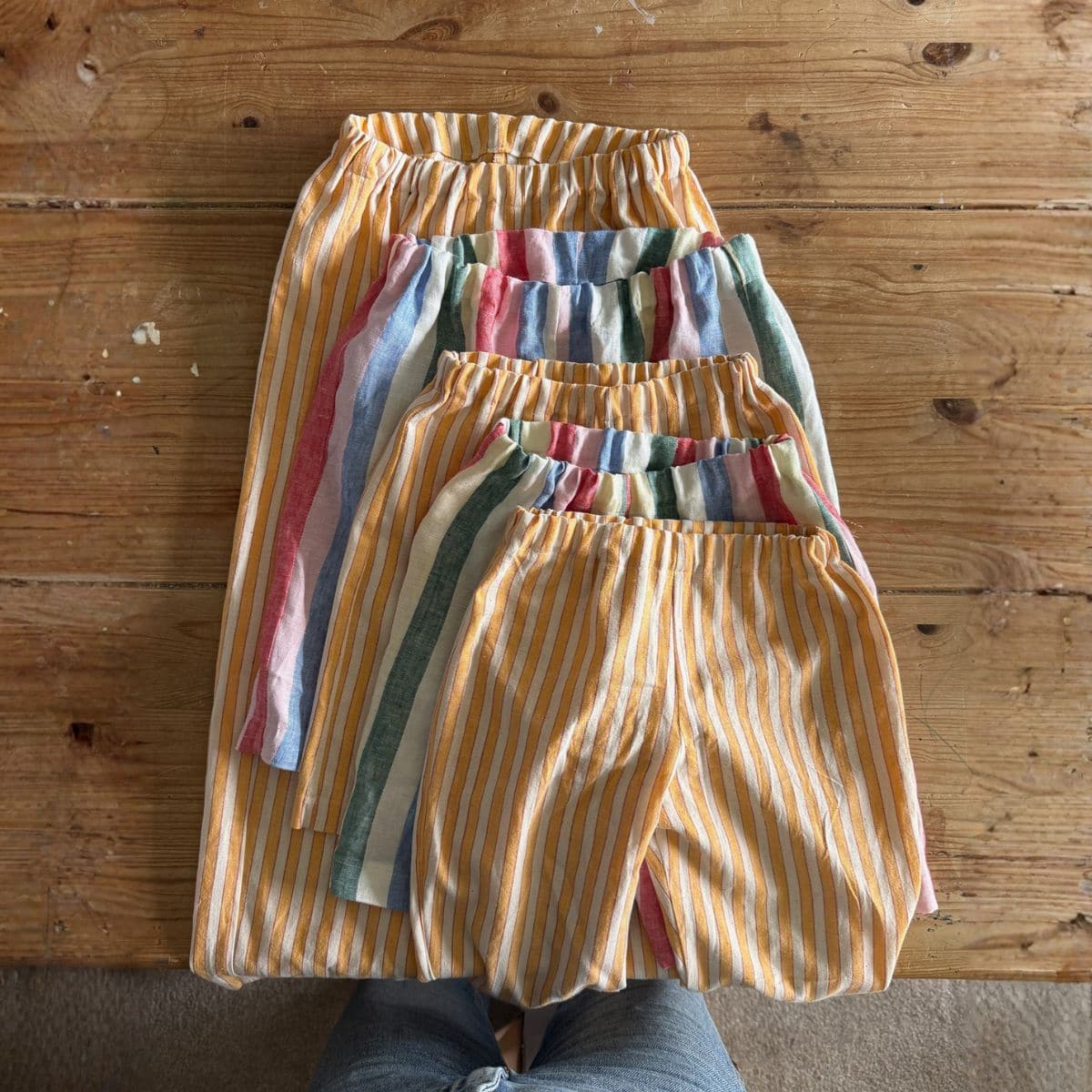 Four pairs of striped shorts in different sizes are arranged on a wooden table, two with yellow and white stripes and two with multi-coloured stripes. Someone wearing jeans is partially visible at the bottom.