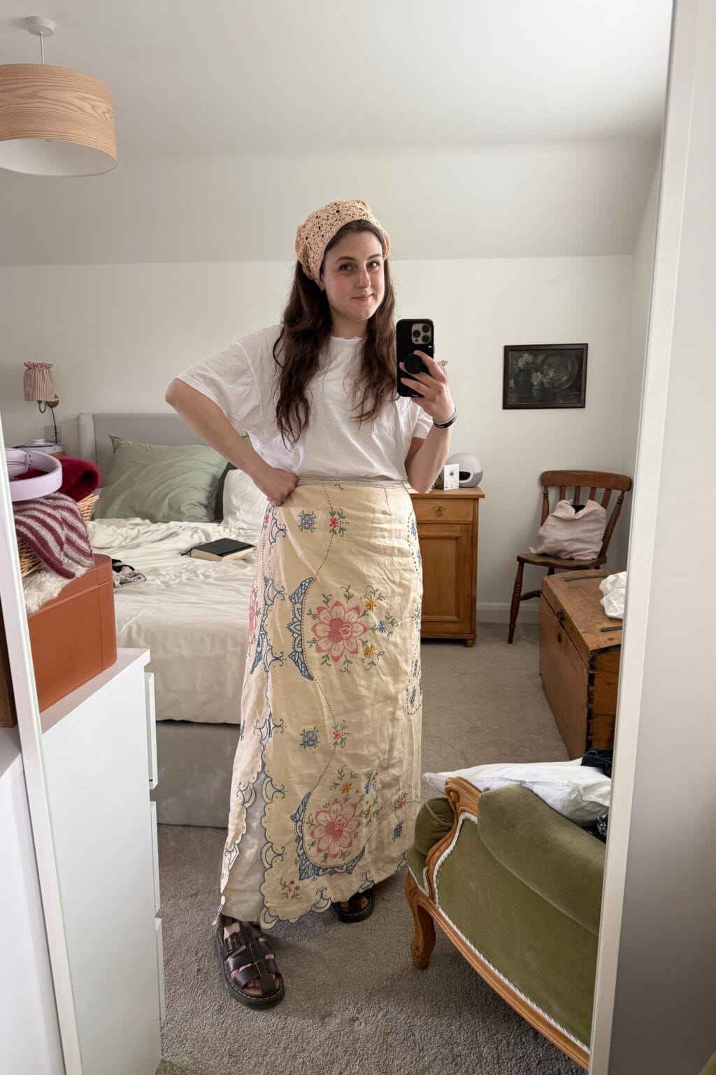 A woman stands in a bedroom taking a mirror selfie, wearing a white T-shirt, beige headscarf, sandals, and a long embroidered skirt—perfect inspiration if you’re wondering how to make a skirt out of a tablecloth. The room is softly lit and inviting.