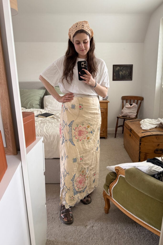 A woman stands in a bedroom taking a mirror selfie. She wears a white shirt, beige floral skirt that looks like it could show how to make a skirt out of a tablecloth, sandals, and a crocheted headscarf. The room has cosy wooden furniture and an armchair.