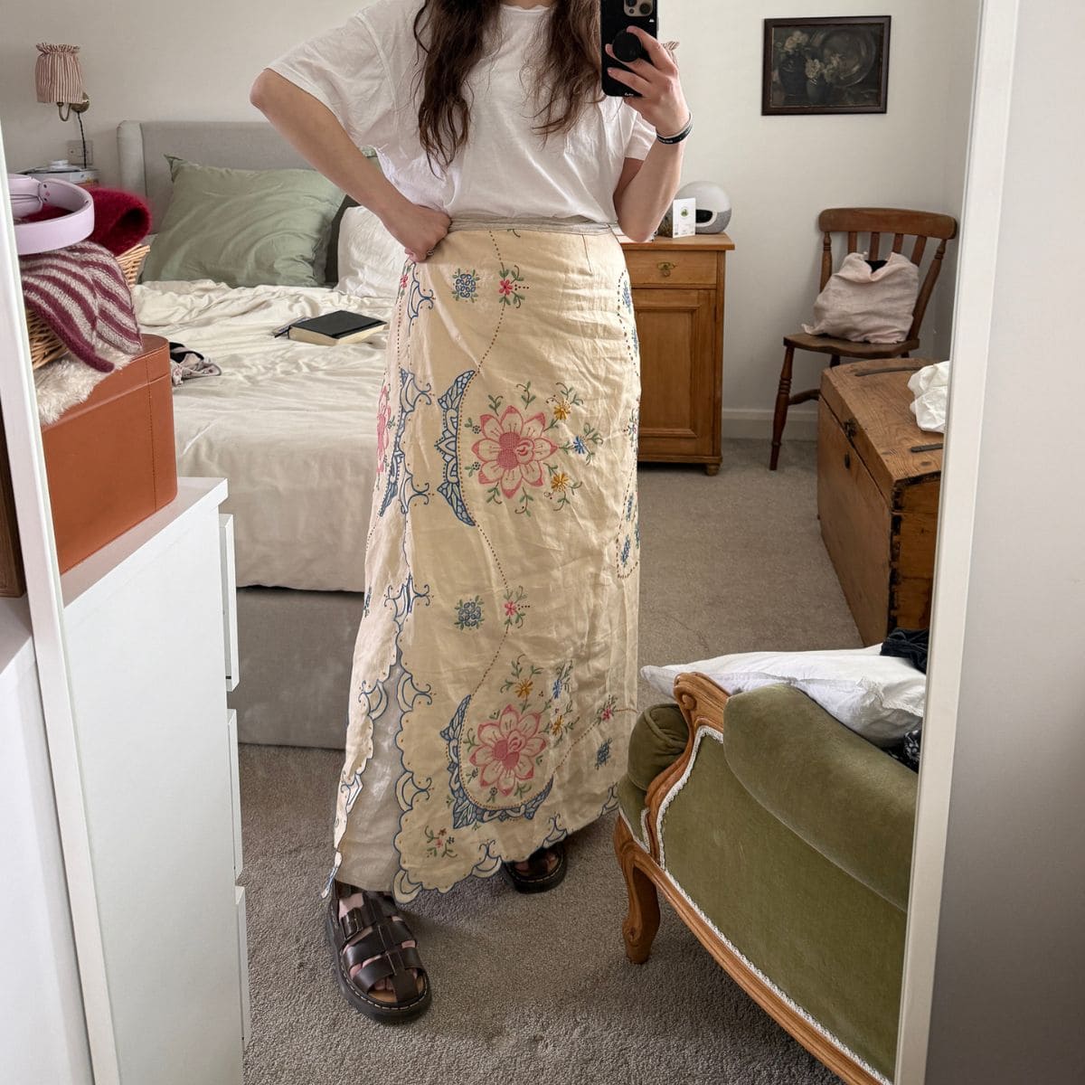 A woman takes a mirror selfie in a bedroom, wearing a loose white T-shirt and a long, cream-coloured skirt with colourful floral embroidery—giving inspiration on how to make a skirt out of a tablecloth—while standing in sandals on a beige carpet near wooden furniture.