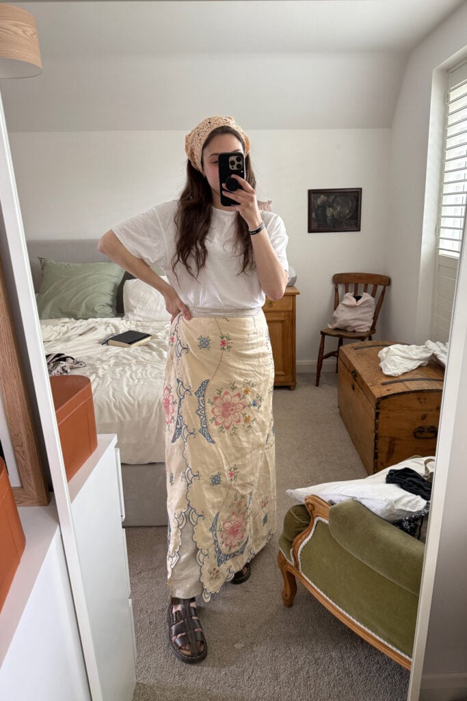 A woman stands in front of a mirror taking a selfie. She wears a white shirt, sandals, headscarf, and a floral skirt—perfect for those curious about how to make a skirt out of a tablecloth. The cosy bedroom features classic decor.