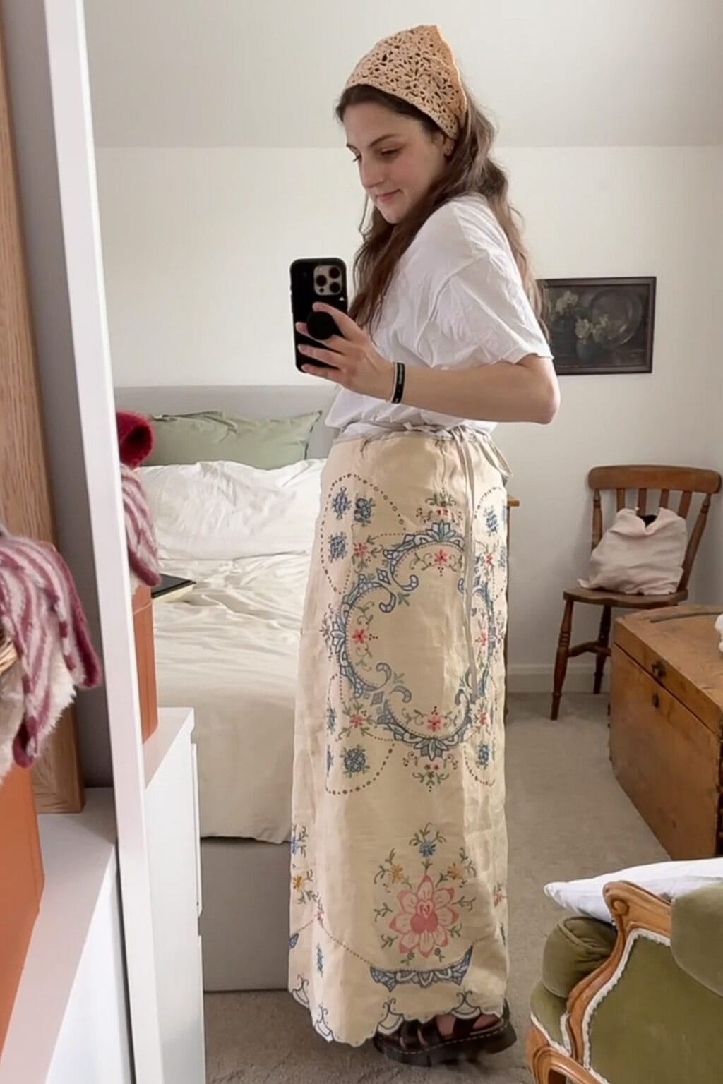 A woman stands in a bedroom taking a mirror selfie. She wears a white shirt, sandals, a beige headscarf, and a unique wrap skirt—perfect inspiration if you’re wondering how to make a skirt out of a tablecloth. The room features a bed, wooden chair, and chest.