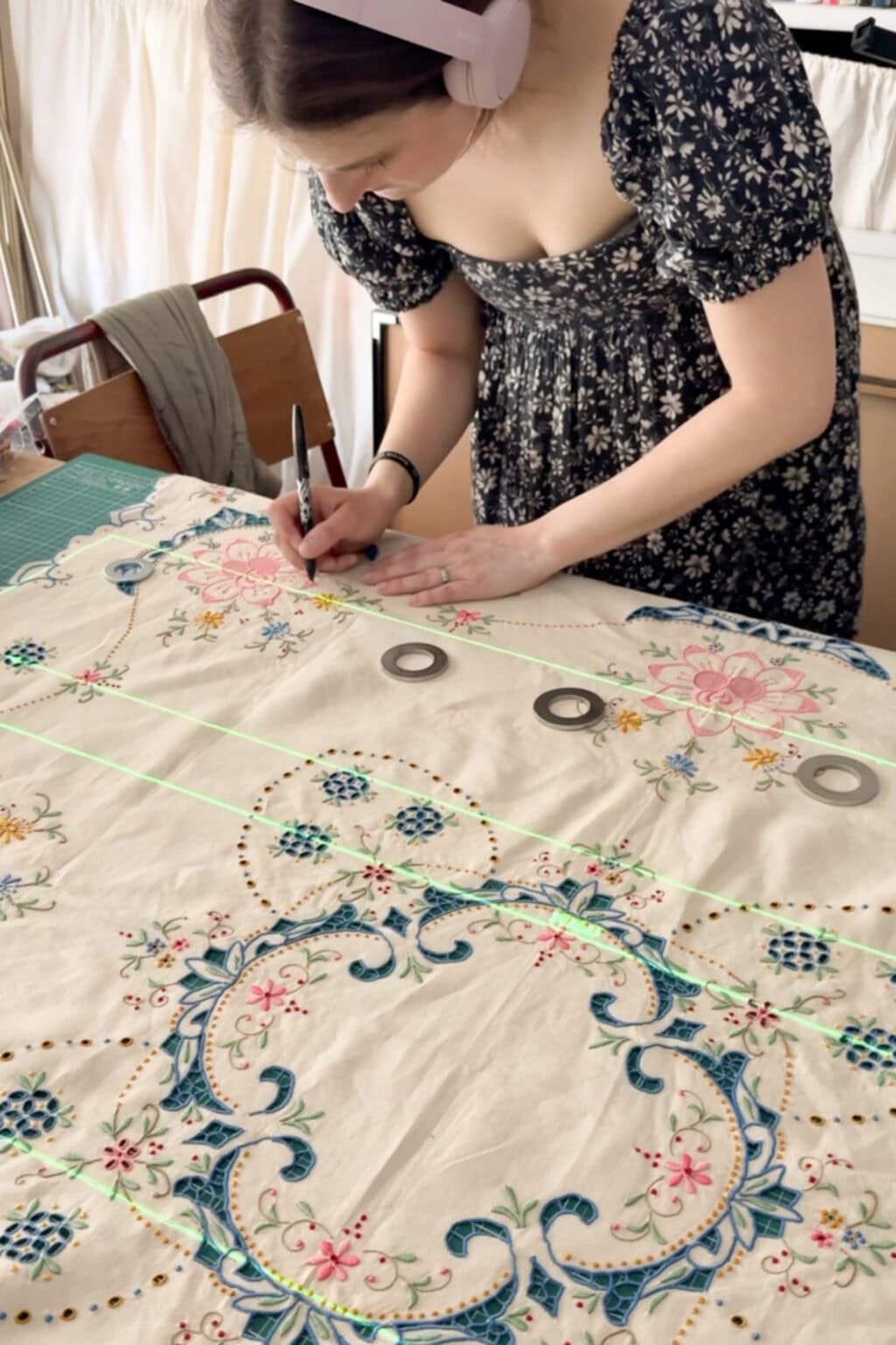 A woman in a black floral dress uses a marker to draw lines on a tablecloth as she works intently at a table in a well-lit room.