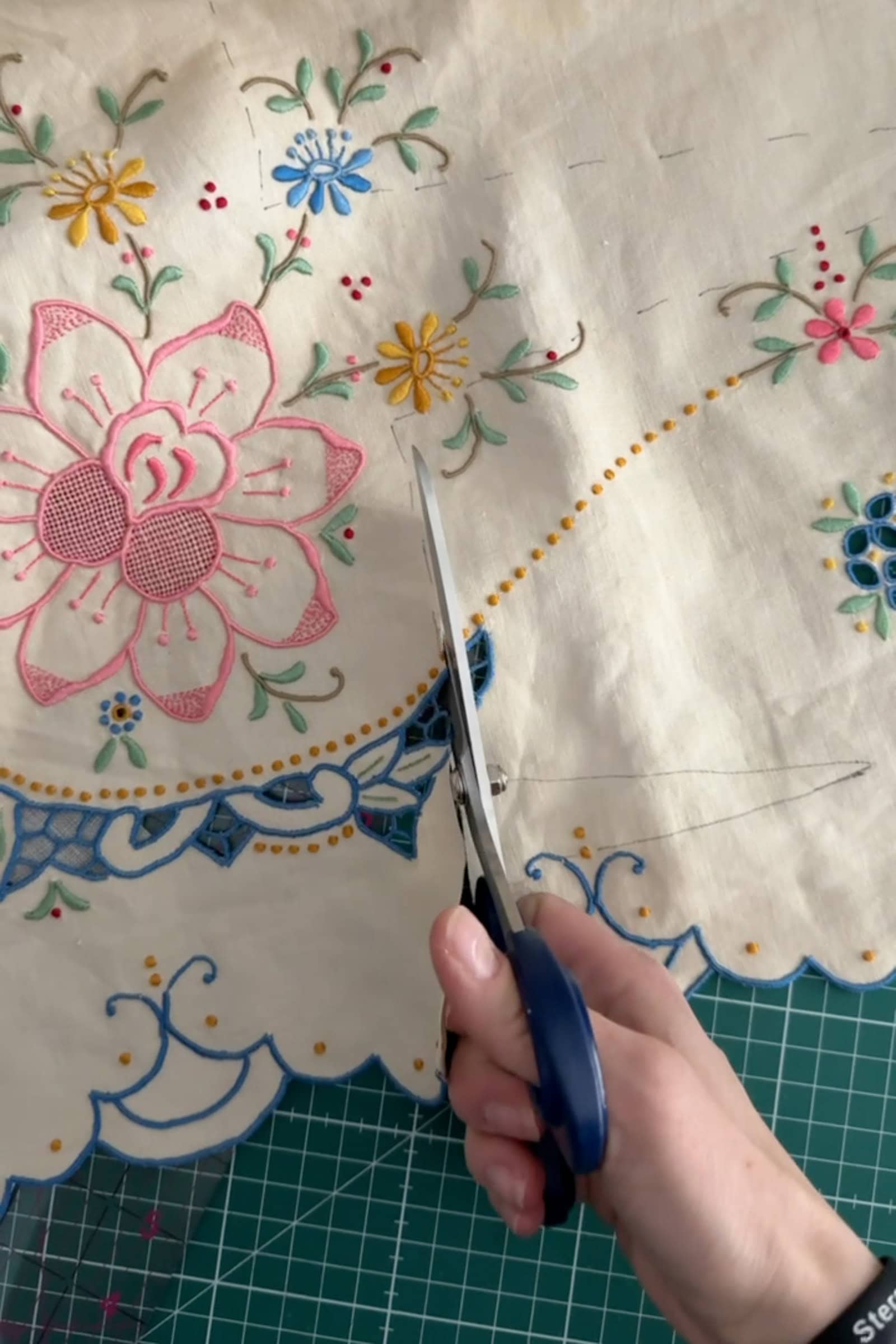 A hand holding scissors cuts into cream-coloured fabric with colourful embroidered floral designs and scalloped blue edges, on top of a green cutting mat.