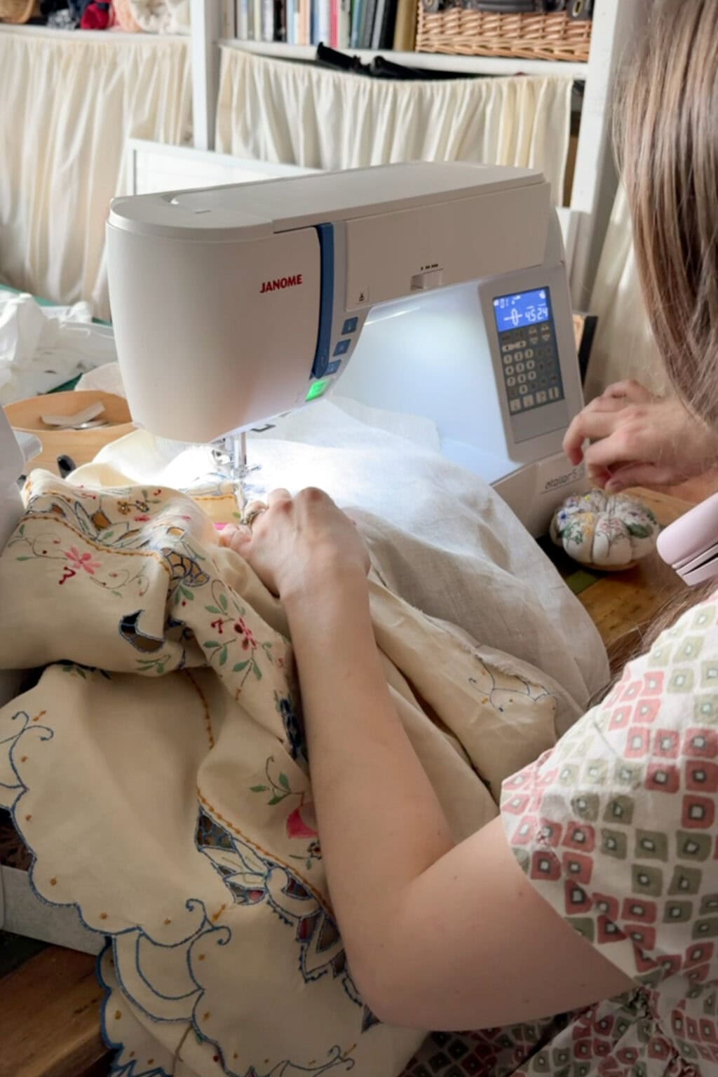 A person uses a sewing machine to stitch patterned fabric. They hold the material with both hands while a pincushion rests nearby, surrounded by shelves filled with books and baskets.