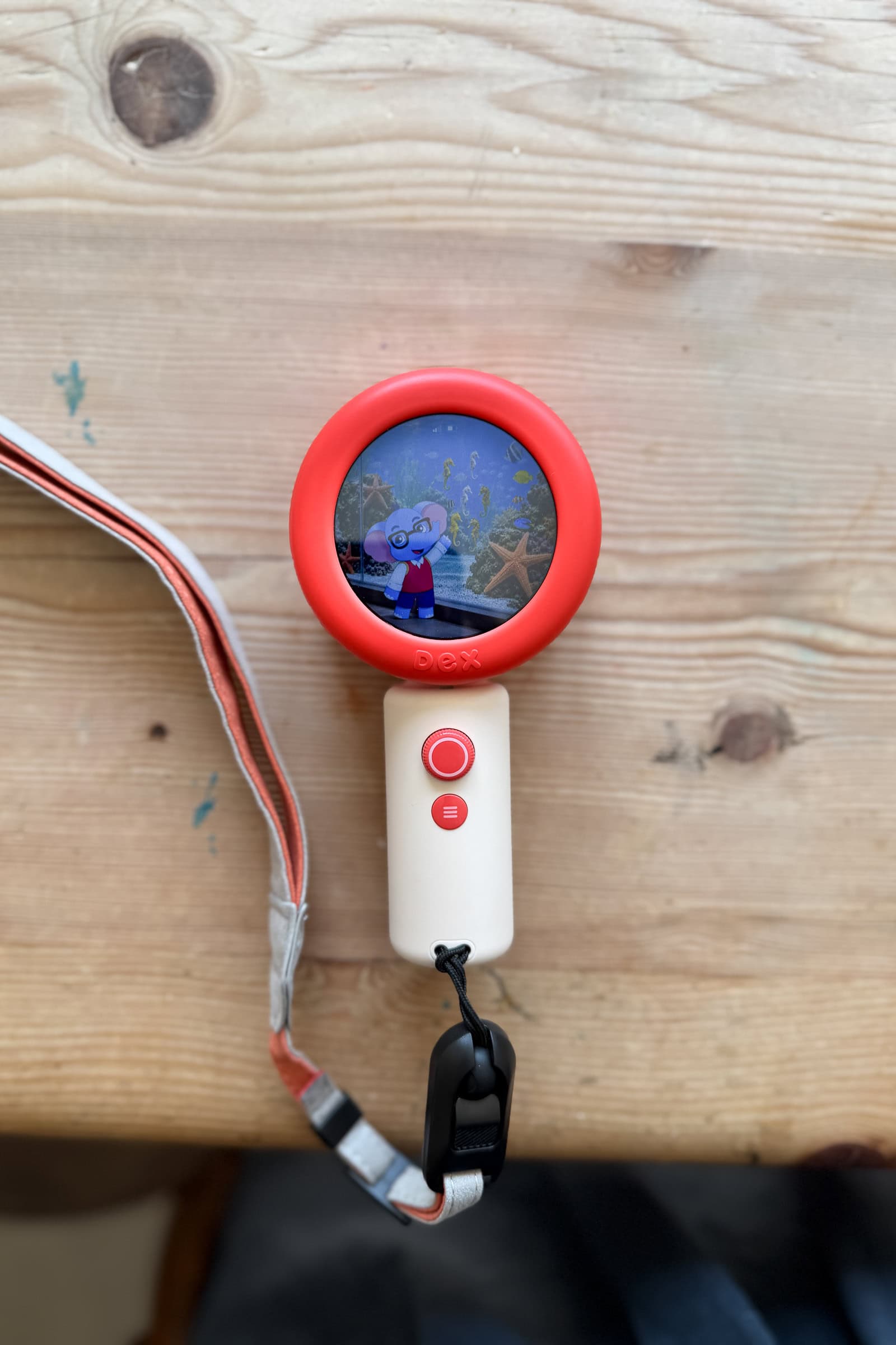 A red and white handheld digital device for children with a wrist strap displays an animated character on its circular screen, ideal for language learning, resting on a light wooden surface.