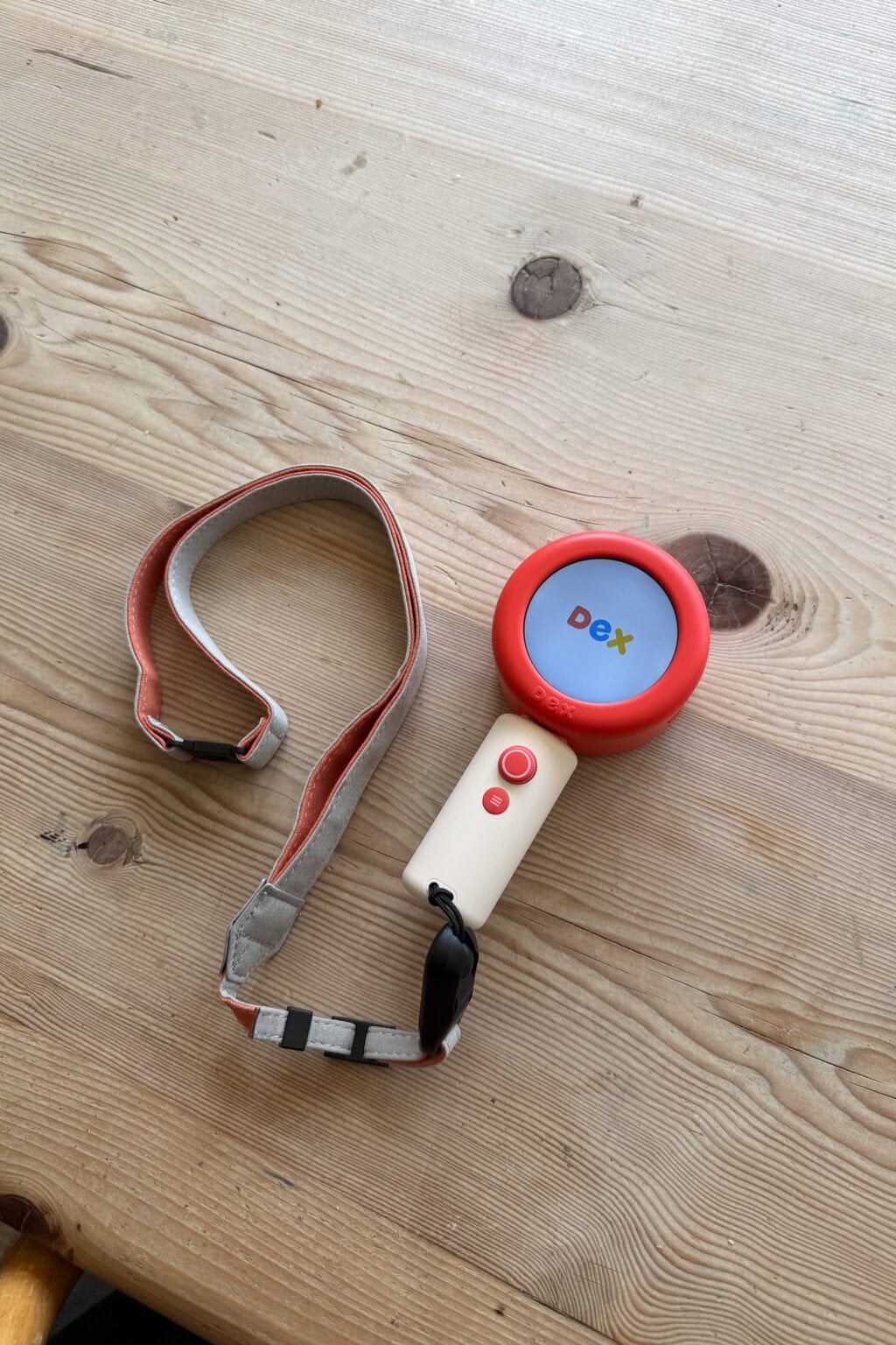 A handheld dex camera for children with a red circular screen displaying the word Dex, attached to a beige remote and a grey-and-red lanyard, rests on a light wooden table—ideal for language learning 