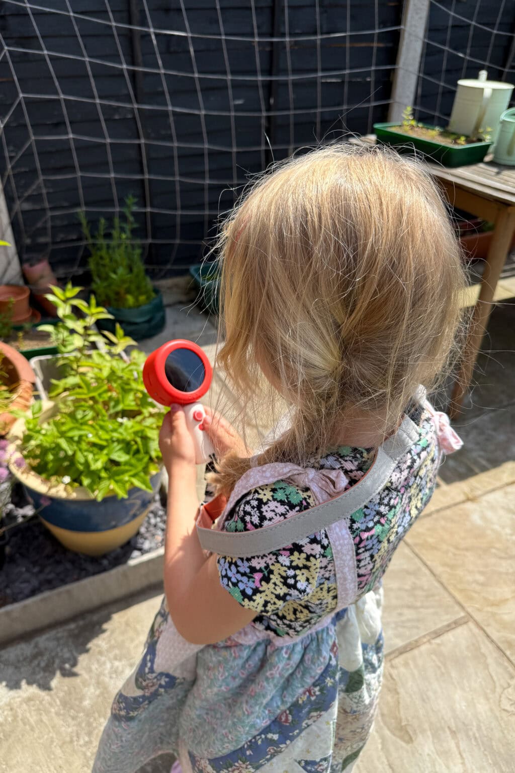 A young girl with blonde hair, wearing a floral dress and apron, holds a red dex camera whilst examining a potted plant outdoors on a sunny day.