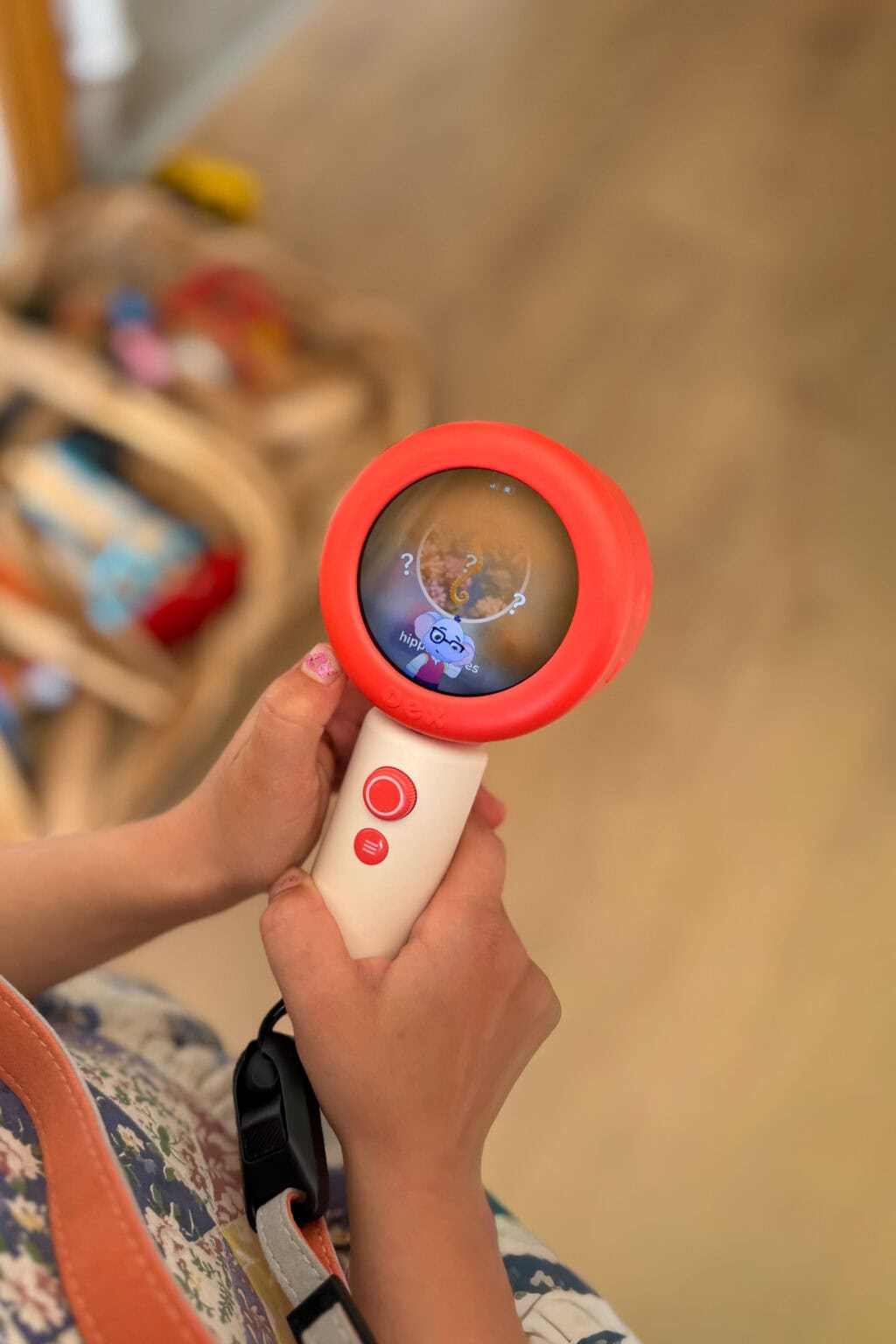 A child holding a red and white dex camera, often used for language learning, displays a cartoon image on its screen. The child has pink nail varnish, wears a patterned dress, and blurry toys are visible in the background.