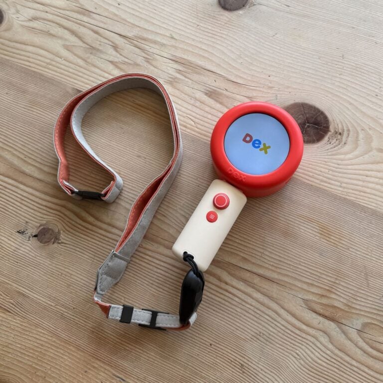 A handheld dex camera with a red circular screen displaying d e x in colourful font, attached to a beige handle with buttons and a grey-and-red adjustable strap—an ideal best tool for language learning for children, pictured on a wooden surface.