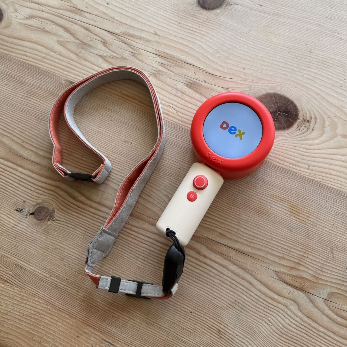 A handheld dex camera with a red circular screen displaying d e x in colourful font, attached to a beige handle with buttons and a grey-and-red adjustable strap—an ideal best tool for language learning for children, pictured on a wooden surface.