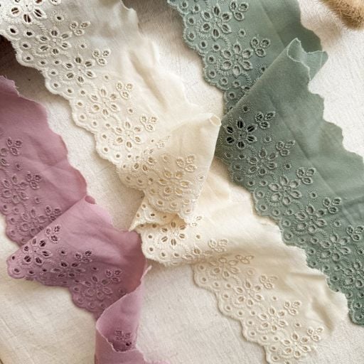Three strips of scalloped broderie anglaise fabric in soft pastel colours—lavender, cream, and sage green—are arranged on a neutral background. Perfect for a beginner sewing project or scrunchie sewing tutorial, each piece features floral embroidery along the edges.