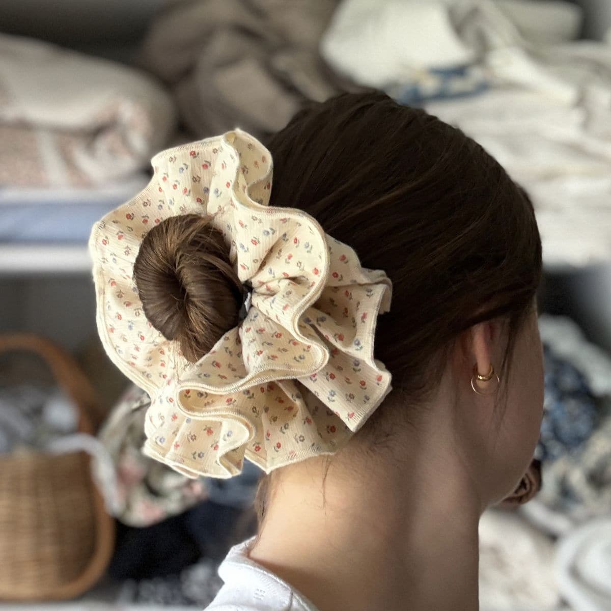 A person with brown hair is wearing a large, cream-coloured floral scrunchie—perfect for a beginner sewing project—in a bun. They have a gold hoop earring and are seen from behind, with blurred fabric and a woven basket in the background.