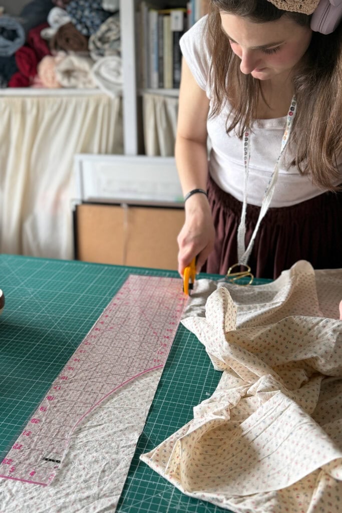 A woman demonstrates a beginner sewing technique, using a rotary cutter to cut patterned fabric for a scrunchie on a green mat, guided by a clear ruler. She has a measuring tape round her neck and sewing supplies in the background.