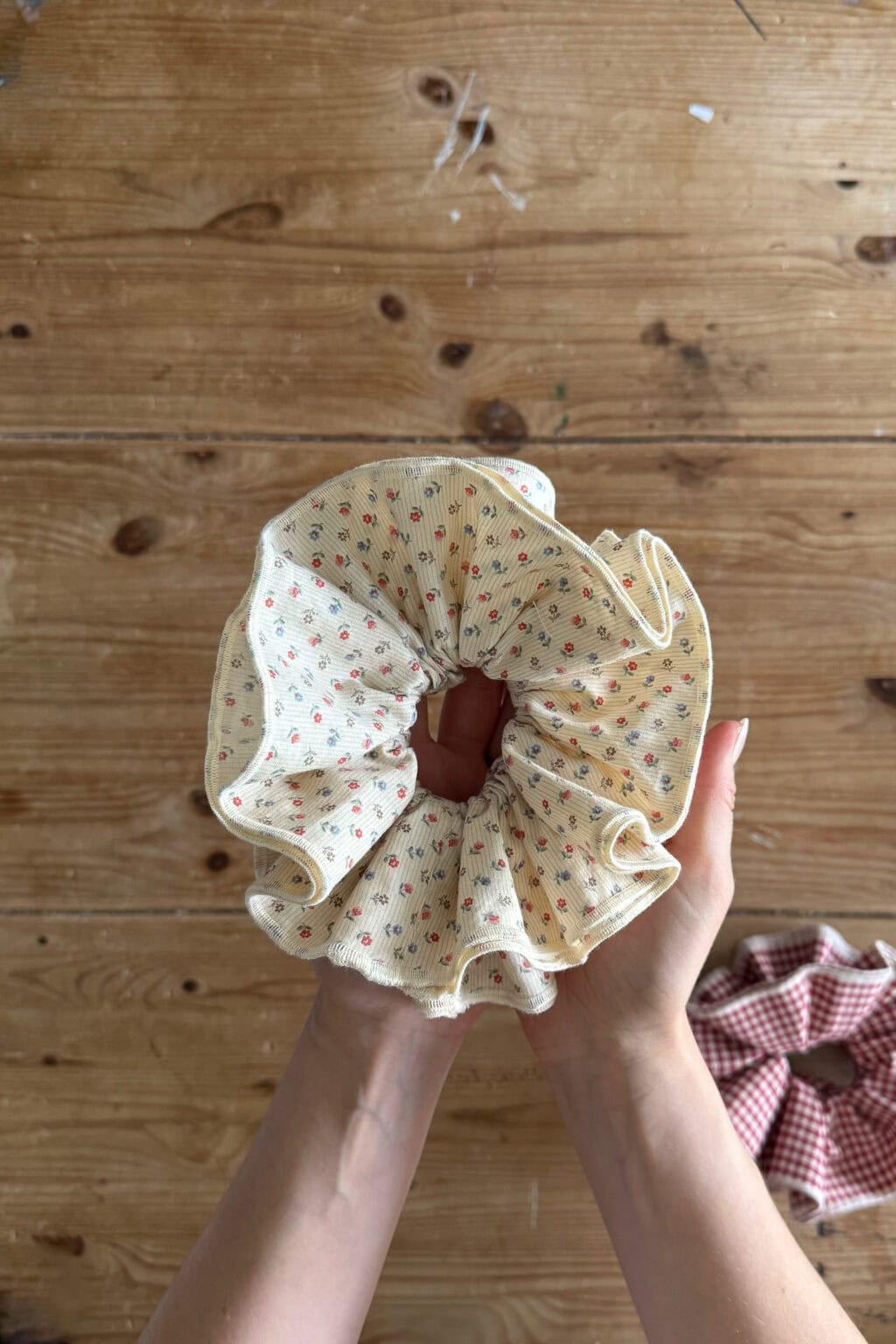 Two hands hold a large, ruffled scrunchie made of cream fabric with small red floral patterns—a fun beginner sewing project—above a wooden table. Another scrunchie with a red and white chequered pattern rests nearby.