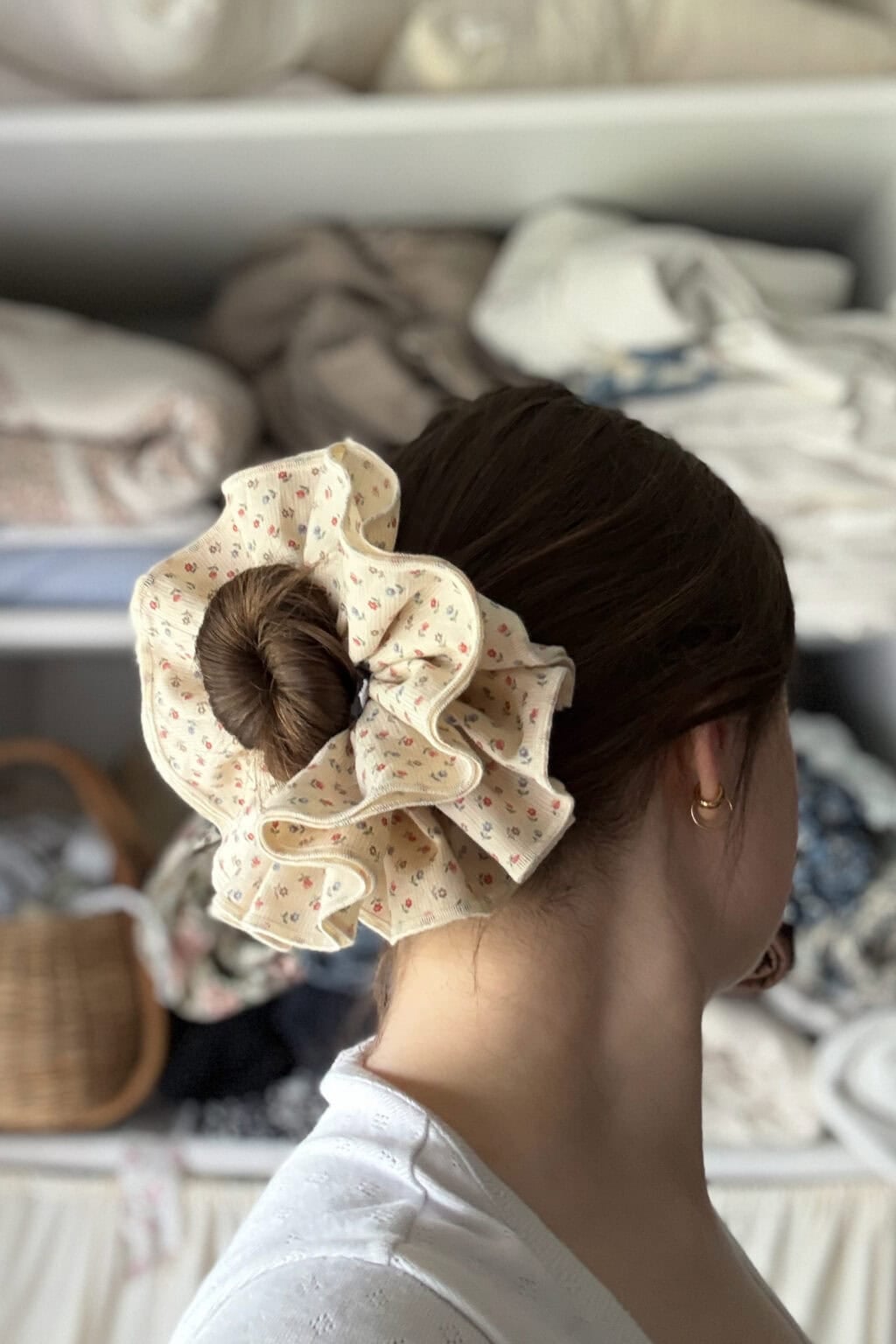 A woman with brown hair in a bun secured by a large, ruffled scrunchie featuring a small floral pattern stands in front of shelves with folded fabrics