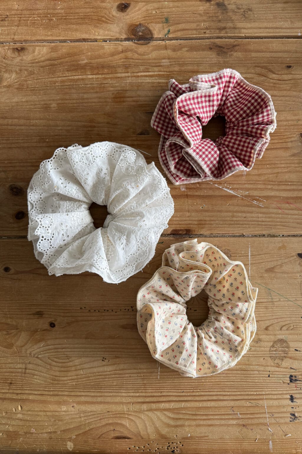 Three scrunchies—one white with lace, one red and white gingham, and one cream with a small floral print—are displayed on a wooden surface, perfect for a beginner sewing project or following a simple sewing tutorial.