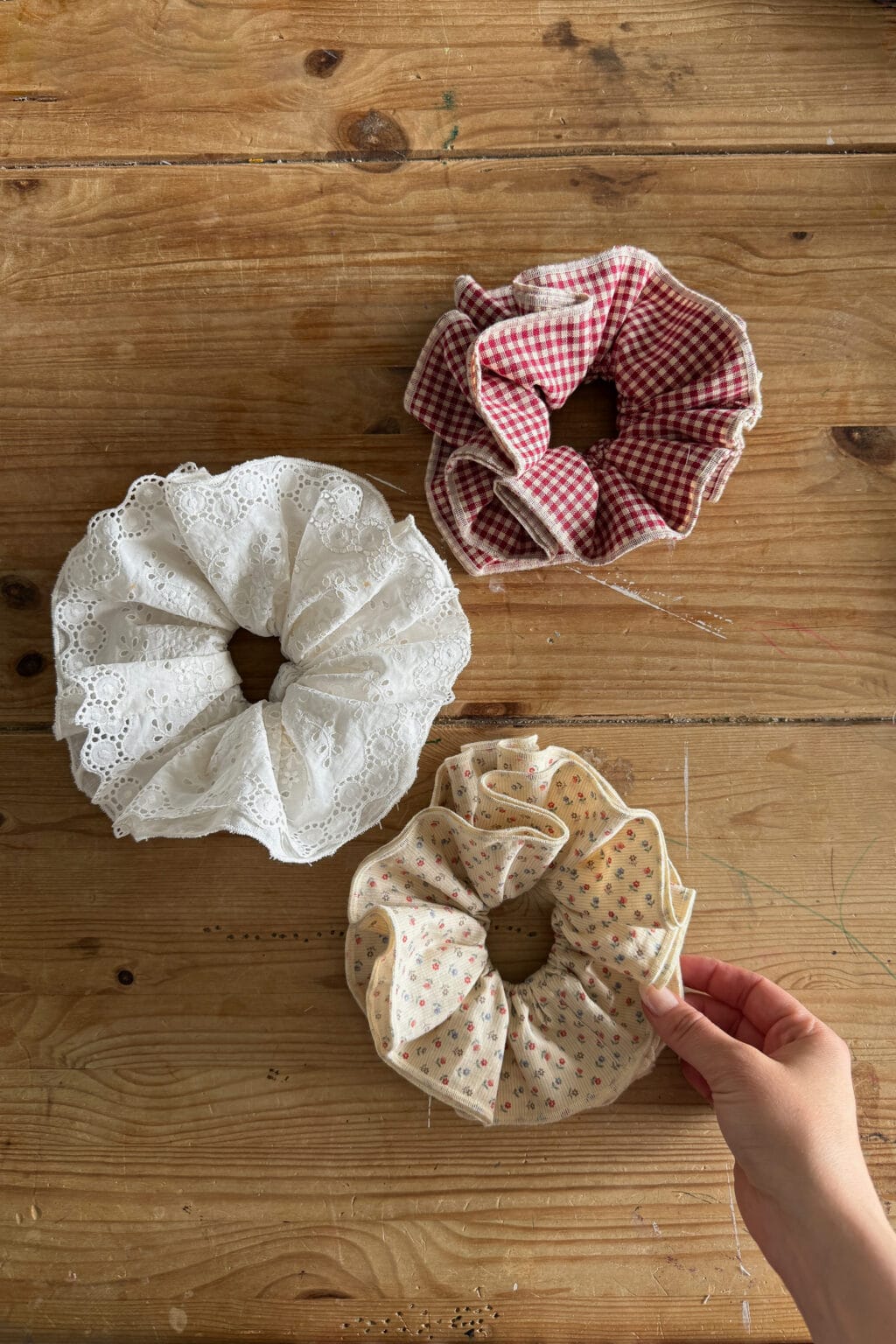 Three large fabric scrunchies rest on a wooden surface: one white with lace, one red and white gingham, and one beige with small floral print, which is being picked up by a hand—perfect inspiration for your next beginner sewing project.