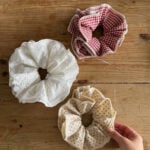 Three large fabric scrunchies rest on a wooden surface: one white with lace, one red and white gingham, and one beige with small floral print, which is being picked up by a hand—perfect inspiration for your next beginner sewing project.