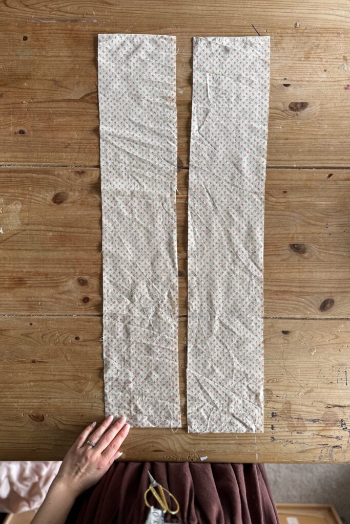Two long, rectangular pieces of patterned fabric are laid side by side on a wooden table. A hand rests at the bottom left corner of the image near the fabric pieces
