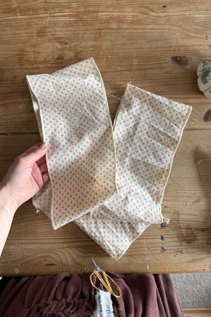 A hand holds two rectangular pieces of fabric with a small floral print, sewn along the edges, on a wooden table