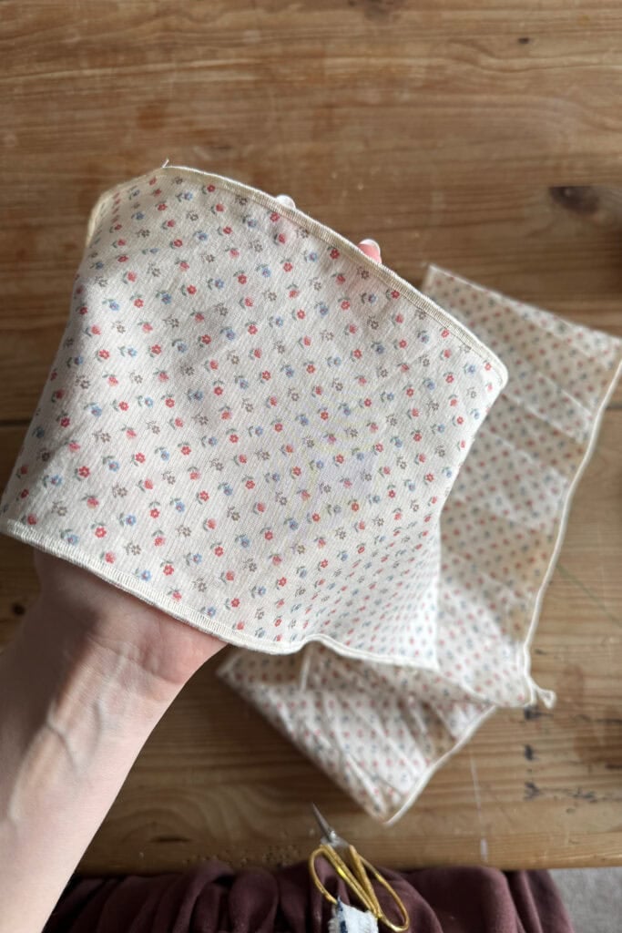 A hand holds a piece of cream-coloured fabric with small pastel floral patterns, ready to make a scrunchie; another matching fabric piece lies on a wooden table in the background.