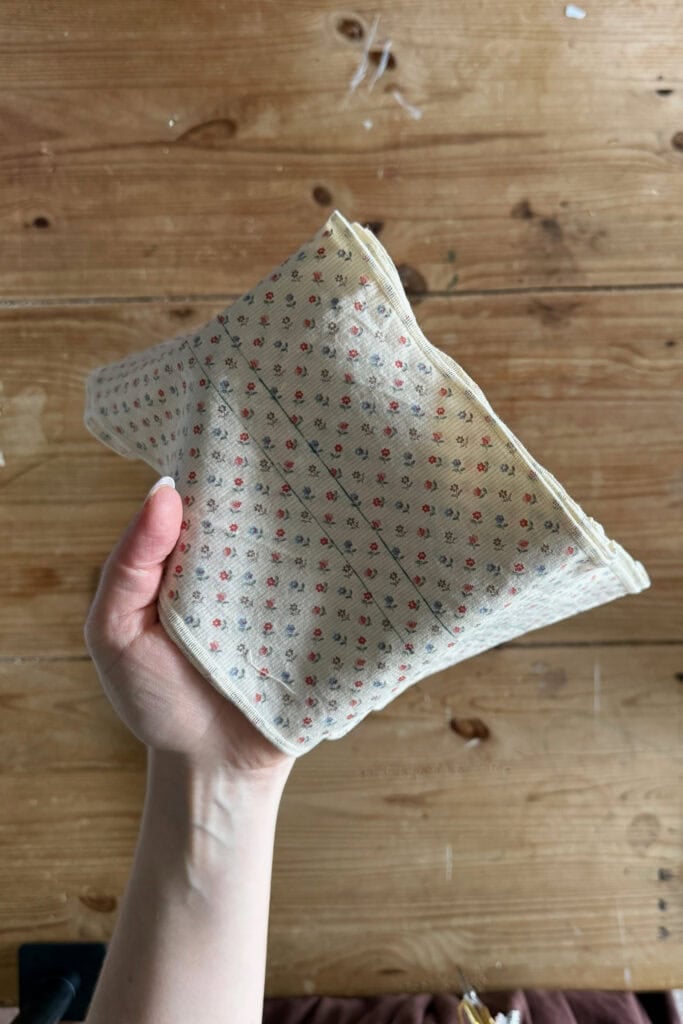 A hand holds a piece of folded cream-coloured fabric with a small red and grey floral pattern, against a wooden surface background.