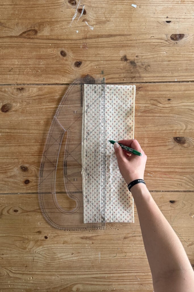 A person’s hand holds a green pen, drawing lines on patterned fabric with a clear plastic ruler, all set on a wooden table