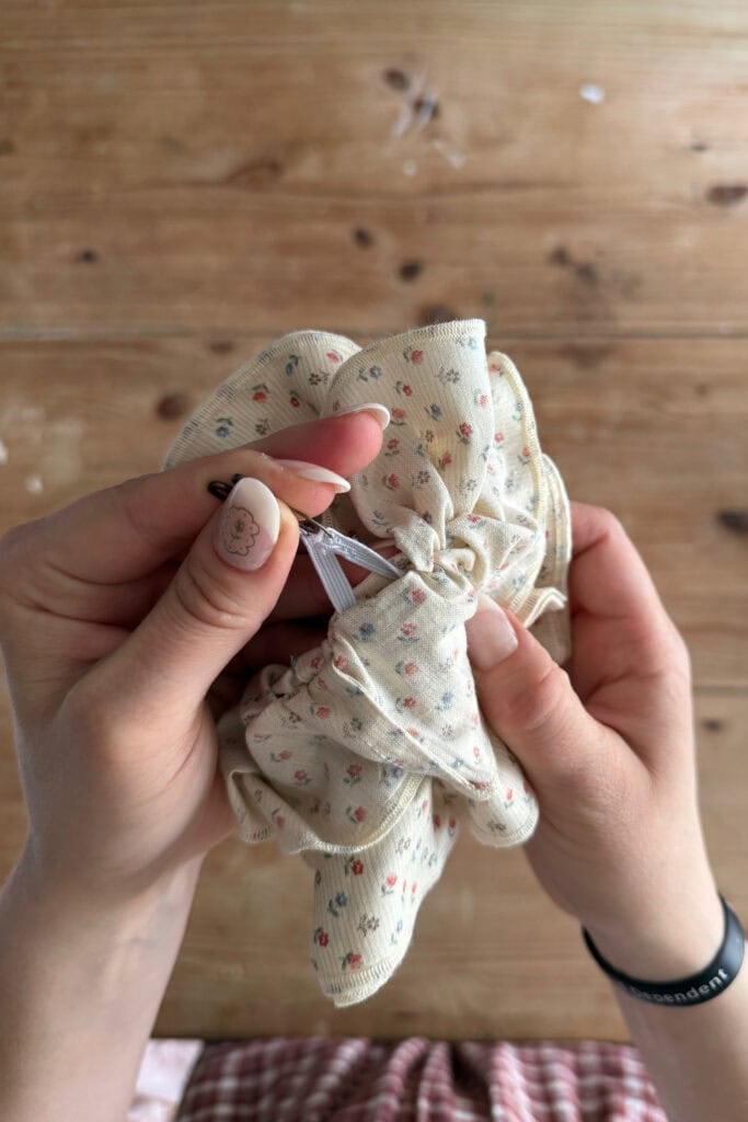 Two hands are creating a floral-patterned scrunchie with a needle and thread, perfect for a beginner sewing project. The person sports a bear design on their thumbnail, wears a black bracelet, and works over a wooden surface.