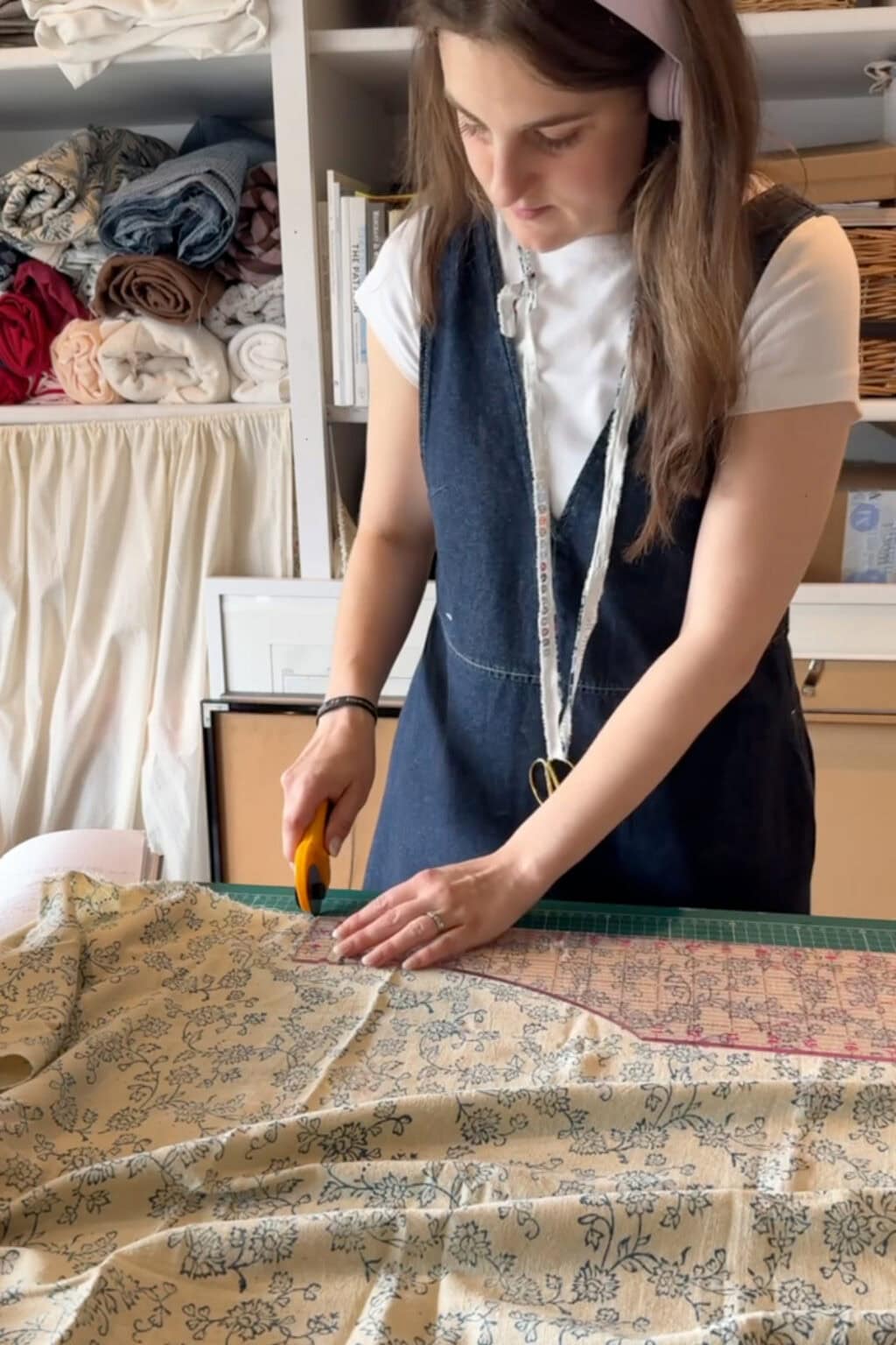 A woman cuts patterned fabric with a rotary cutter on a table, surrounded by shelves with folded fabrics. She wears a white shirt, denim pinafore, and a measuring tape around her neck—capturing the perfect moment for anyone starting sewing or looking for inspiration in this beginner’s guide to making clothes.