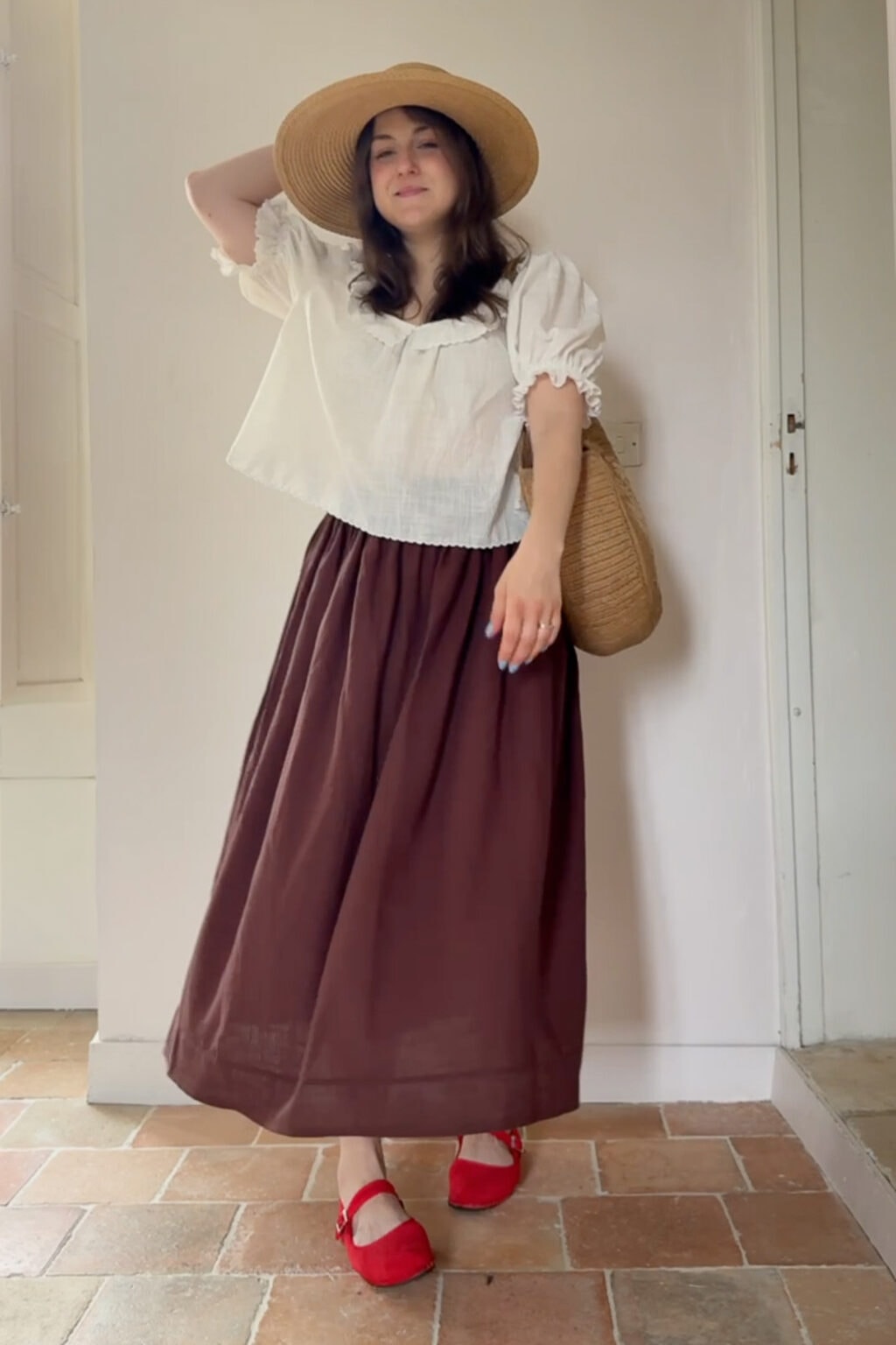 A woman wearing a wide-brimmed hat, white blouse, long brown skirt, red shoes, and a woven bag stands indoors on a tiled floor, posing with one arm raised—a graceful look that could easily inspire anyone eager to sew clothes for themselves 