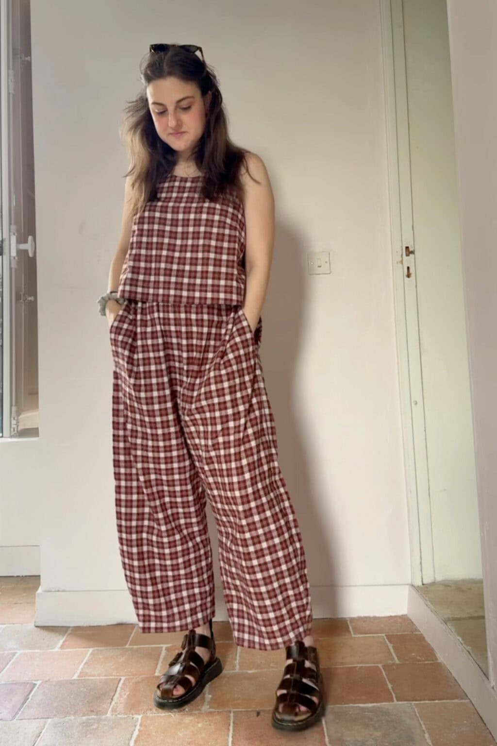 A person stands indoors wearing a sleeveless, red and white chequered jumpsuit—an ideal project for those eager to sew clothes—with hands in pockets, dark sandals, and sunglasses resting on their head. They are looking down with a relaxed expression.
