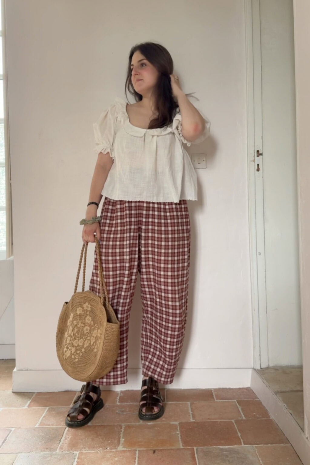 A woman stands indoors wearing a white blouse, red checked trousers, and black sandals, holding a large straw bag with a floral pattern. Perfect for those interested in starting sewing or looking for inspiration to sew clothes, her look combines classic style with creative flair. She has dark hair and looks to the side, touching her hair.
