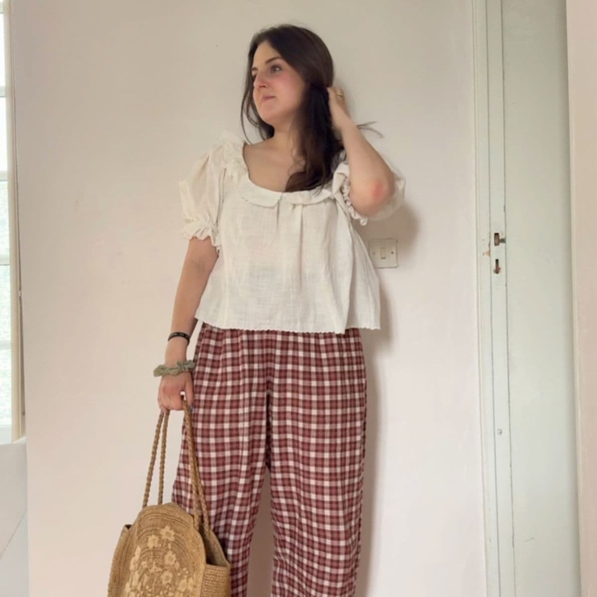 Woman stands indoors by a white wall, wearing a white blouse with puff sleeves and loose red checked trousers. Perfect for those starting sewing, her outfit offers inspiration to sew clothes that are both stylish and comfortable. She holds a woven straw bag and touches her hair, looking slightly to the side.